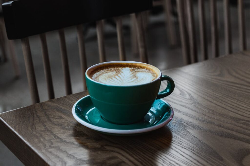 A beautifully crafted latte with leaf art in a green cup on a wooden table inside a cafe.
